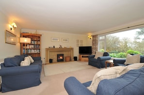 A cozy living room with a fireplace, a bookshelf, a TV, and a large window offering a view of greenery. - Tregillan (Wadebridge)