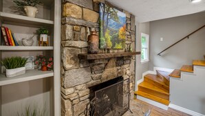 Una chimenea de piedra con un cuadro encima, escaleras de madera, y estantes con libros y plantas.