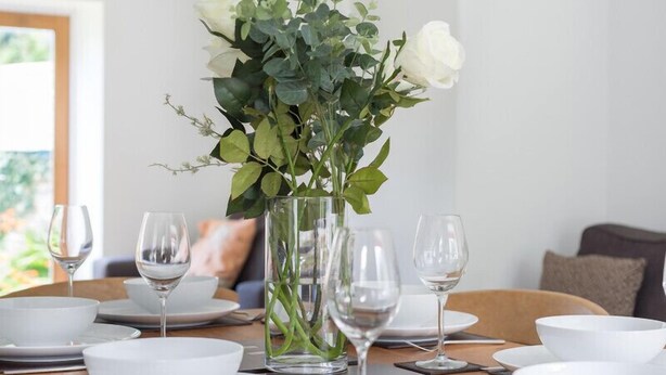Une table à manger dressée avec de l’assiette blanche, des bols et des verres à vin, agrémentée d’un vase rempli de fleurs blanches et de feuillage vert.
