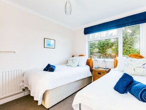 A double room with two beds, a window with a view of greenery, a radiator, and a framed picture on the wall. - Little Sailing (Wadebridge)