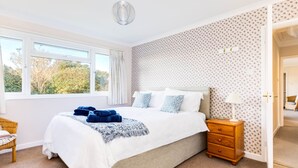 A bedroom with a large bed, a bedside table, a lamp, and a window with curtains. - Little Sailing (Wadebridge)