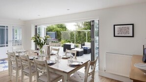 A dining area with a wooden table set for six, white chairs, and a view of the garden through glass doors. - Cherrytrees (Wadebridge)
