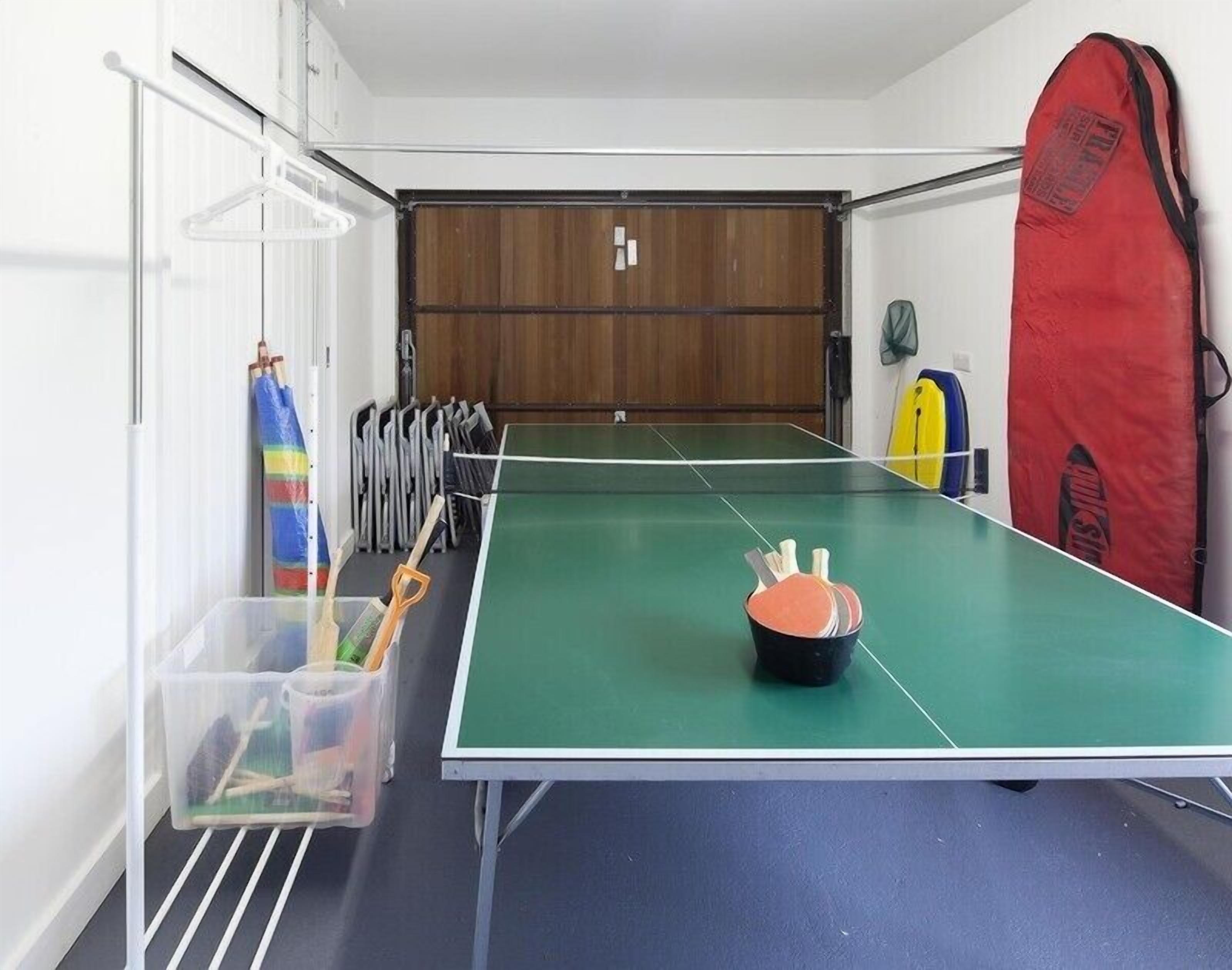 A ping pong table in a room with a wooden door, a red paddle bag, and a plastic storage bin with sports equipment.