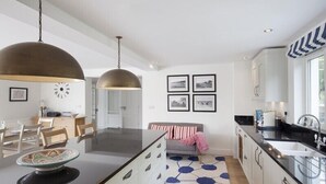 A modern kitchen with a central island, pendant lighting, and a dining area with a clock and framed pictures on the wall. - Cherrytrees (Wadebridge)