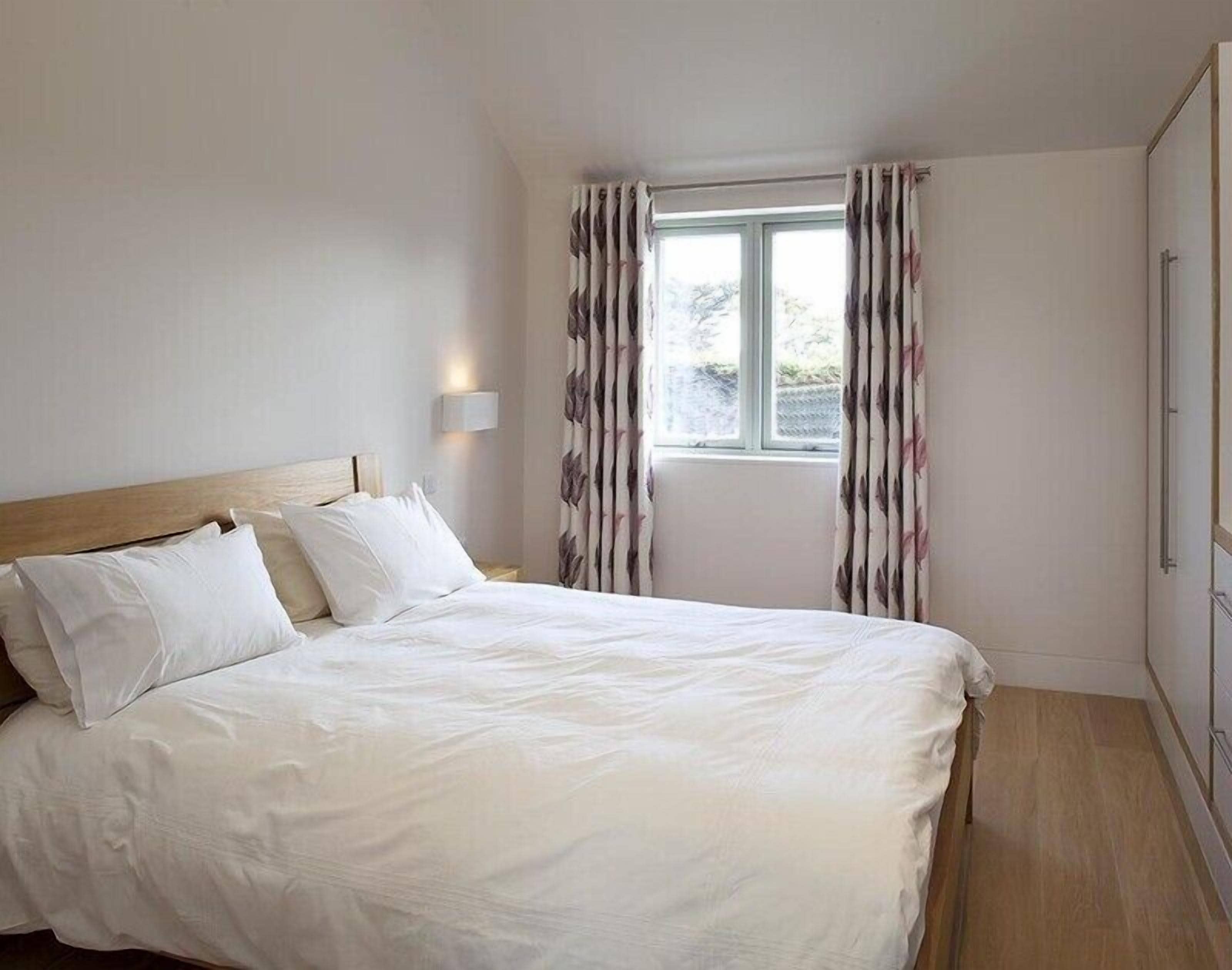 Cama bem arrumada com lençóis brancos em quarto com cabeceira de madeira, janela com cortinas florais e luminária fixa na parede.