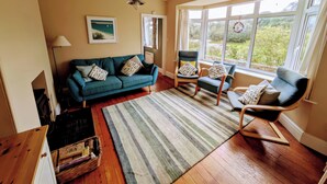 A living room with a teal sofa, two armchairs, a striped rug, and a large window with a view of greenery.