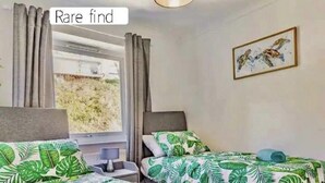 A twin room with two beds, a window with curtains, a bedside table with a lamp, and a painting on the wall. - Lovely Apartment Sleeps 6 Singles in Torquay (Torquay)