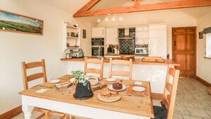 A dining area with a wooden table and chairs, a kitchen in the background, and a painting on the wall. - Old Hall Byre (Leyburn)