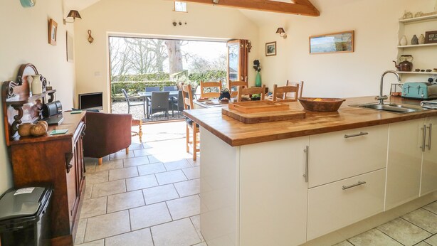 A spacious kitchen with wooden beams, a large island, and a view to the outdoor patio. - Old Hall Byre (Leyburn)