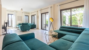 Un vaste espace de vie comprenant un grand canapé vert, un fauteuil vert plus petit et un lampadaire. Une porte-fenêtre coulissante donne accès à un balcon aménagé avec du mobilier d’extérieur.