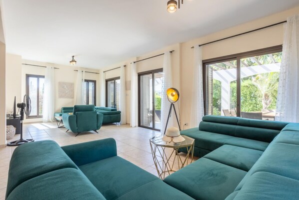 Un vaste espace de vie comprenant un grand canapé vert, un fauteuil vert plus petit et un lampadaire. Une porte-fenêtre coulissante donne accès à un balcon aménagé avec du mobilier d’extérieur.