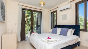 Une chambre avec un grand lit, une table de chevet en bois, un luminaire au plafond et une vue sur les palmiers par les fenêtres.