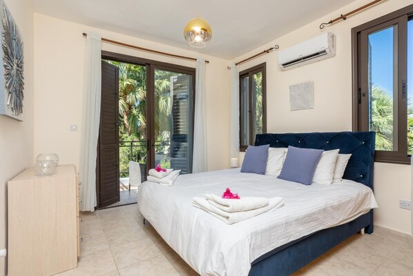 A bedroom with a large bed, a wooden nightstand, a ceiling light, and a view of palm trees through the windows. - Villa Anna - Four Bedroom Villa, Sleeps 8 (Latchi)