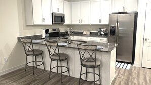 Una cocina con armarios blancos, un refrigerador de acero inoxidable, un microondas y una estufa a gas. Hay tres taburetes de bar con estructura metálica y asientos acolchados. El piso es de madera y la cocina tiene un salpicadero de color claro.