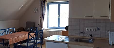 Een kleine keuken met een koelkast, spoelbak en kastjes. Er is een eettafel met stoelen en een raam met gordijnen.