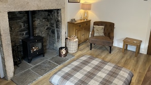 A cozy living room with a stone fireplace, a wooden cabinet, a cushioned armchair, a small table, and a plaid throw blanket. - Bracken Cottage (Bishop Auckland)