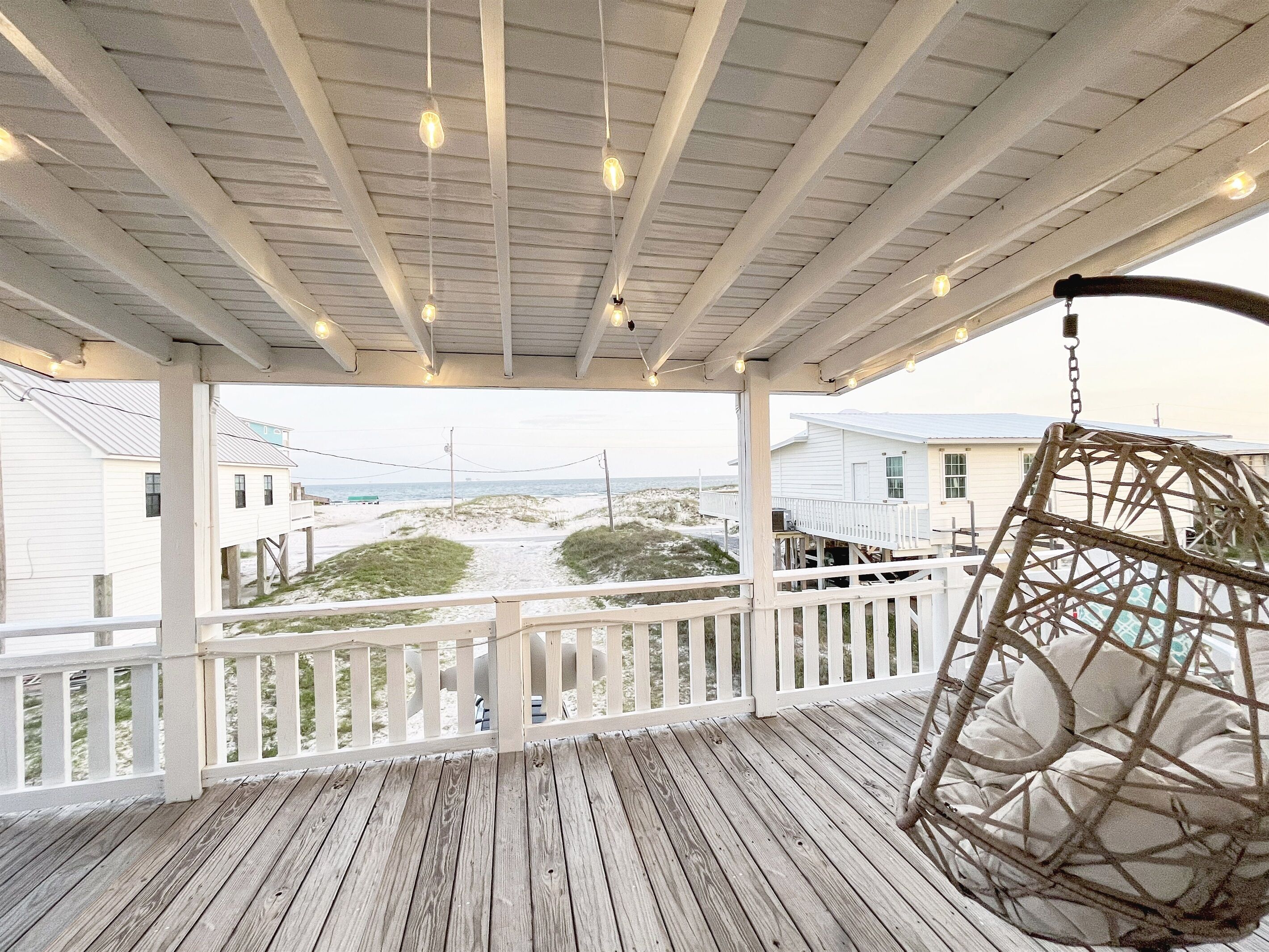 Une terrasse en bois avec une balustrade blanche, une balançoire suspendue et une vue sur la plage et les bâtiments en arrière-plan.