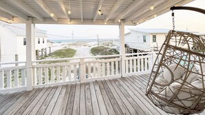 Une terrasse en bois avec une balustrade blanche, une balançoire suspendue et une vue sur la plage et les bâtiments en arrière-plan.