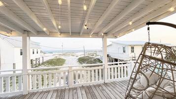 Une terrasse en bois avec une balustrade blanche, une balançoire suspendue et une vue sur la plage et les bâtiments en arrière-plan.
