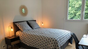 A bedroom with a patterned bedspread, two bedside lamps, a small nightstand, a chair, and a window with a view of trees. - 75 m2 # logia # parking # BESTOFBLO 4 # INSEAD (Fontainebleau)