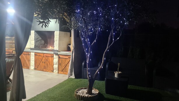 Uma área de pátio ao ar livre com lareira de pedra, móveis de madeira e uma árvore decorada com luzes.