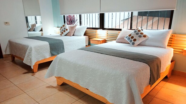 Dos camas individuales con cabeceros de madera y ropa blanca, una ventana con persianas y suelo de baldosas.
