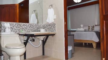A bathroom with a toilet, sink, and mirror. There is a bed with a white blanket and pillows in the adjacent room.