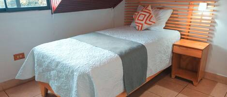 Una cama individual con cabecera de madera, una mesita de noche y una ventana con vista a árboles.