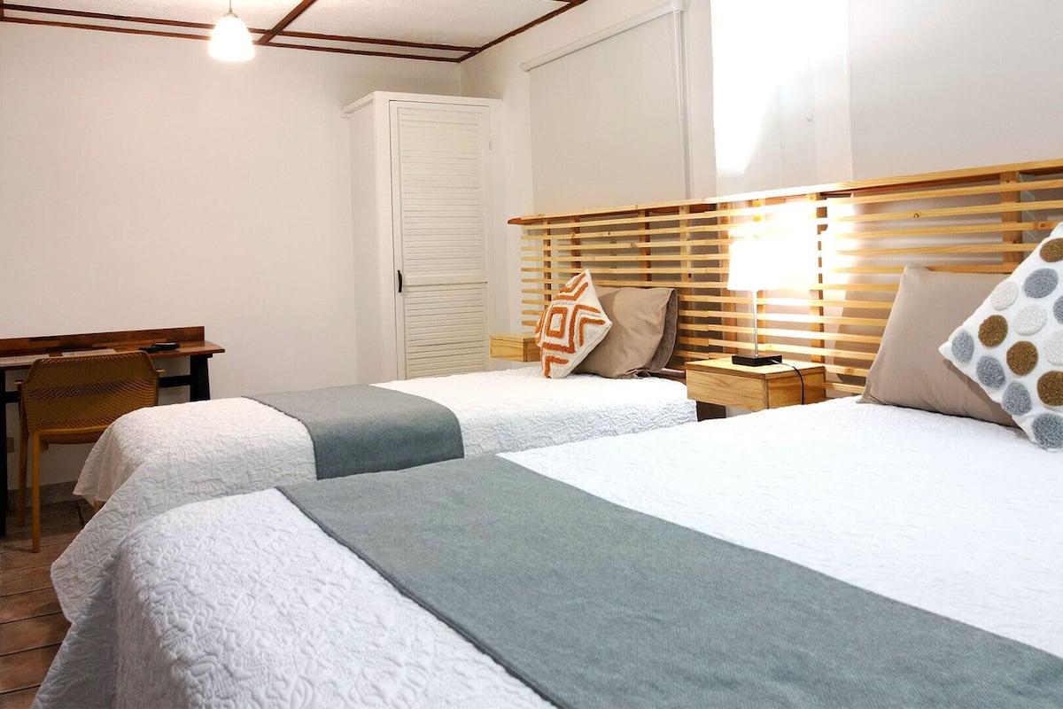 A hotel room with two beds, a desk, a chair, a wardrobe, and a bedside table with a lamp.