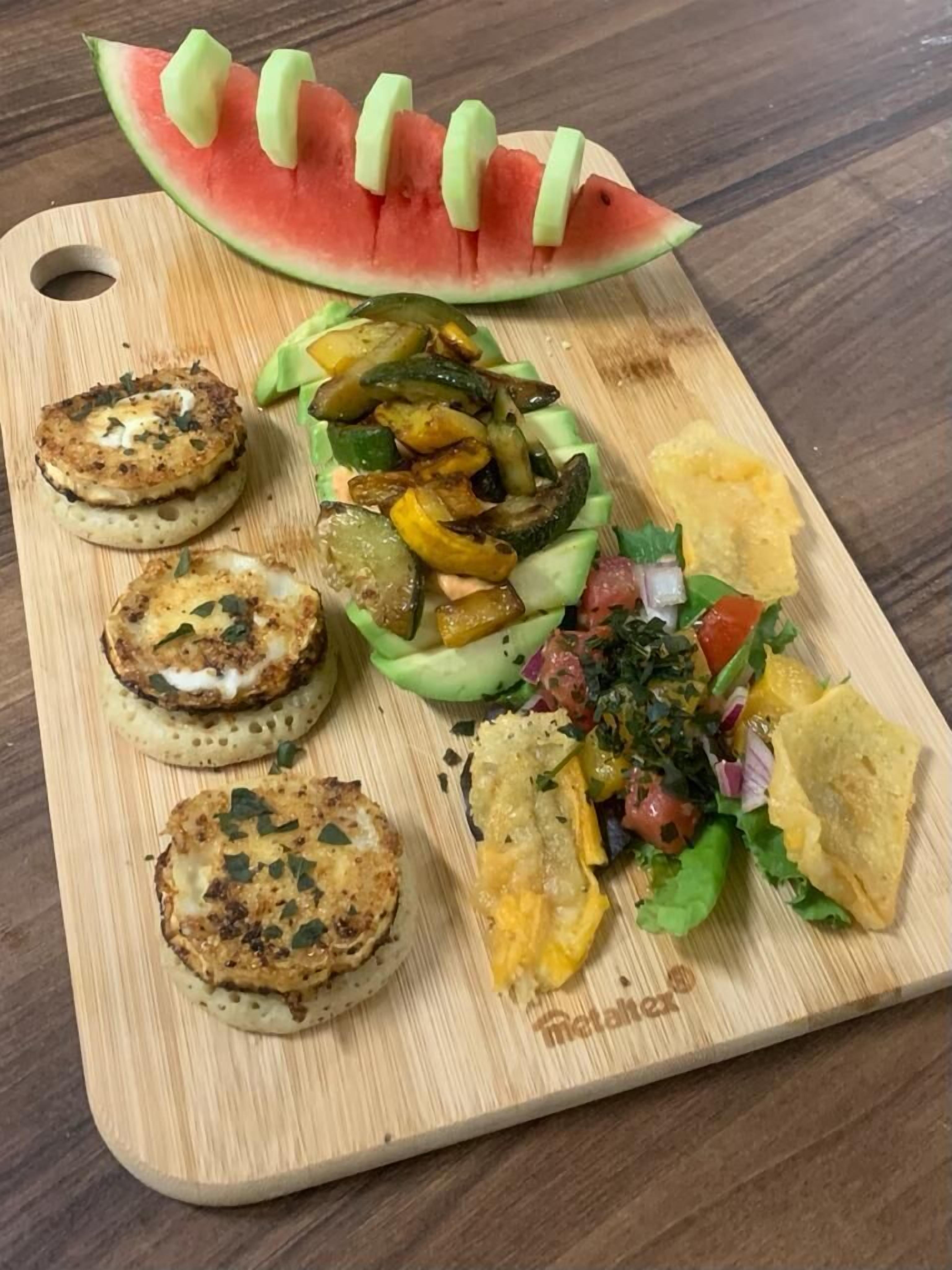 A wooden cutting board with various food items including grilled patties, sliced vegetables, a watermelon slice, and a salad.