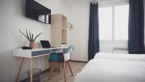 Een hotelkamer met een bed, een bureau met een laptop, een stoel, een kamerplant, een kast en een raam met gordijnen.