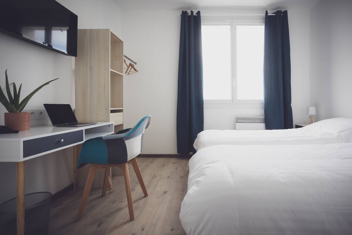 Een hotelkamer met een bureau, laptop, stoel, twee bedden en een raam met gordijnen.