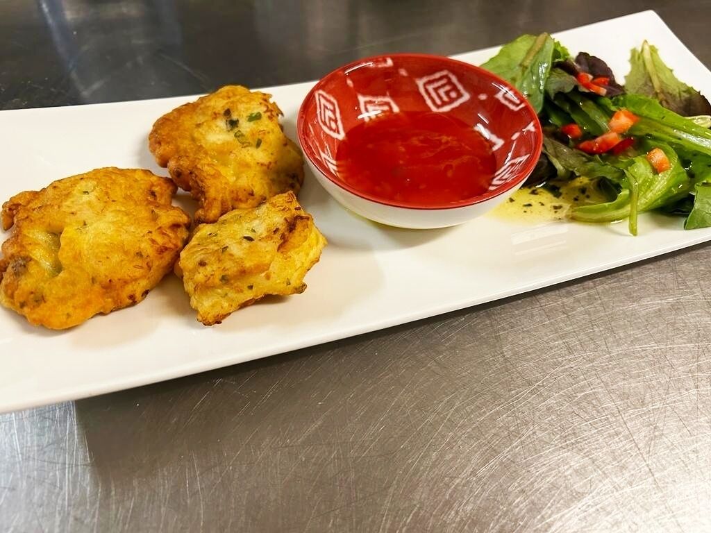 Un plato con tres alimentos fritos y un recipiente de salsa roja, acompañado de una ensalada verde con pimientos rojos.
