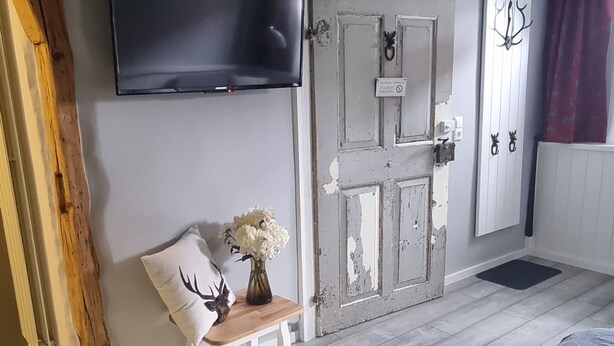 A bedroom with a bed, a television mounted on the wall, a wooden bench with a vase of flowers, and a rustic door. - Lovingly furnished FeWo zum Hirschen (Herzberg am Harz)