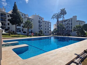 Une piscine à l’eau d’un bleu limpide, entourée d’une terrasse carrelée, et jouxtant un bâtiment doté de nombreux balcons et de palmiers.