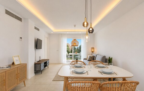 A modern dining area with a white table set for four, wicker chairs, and pendant lighting.