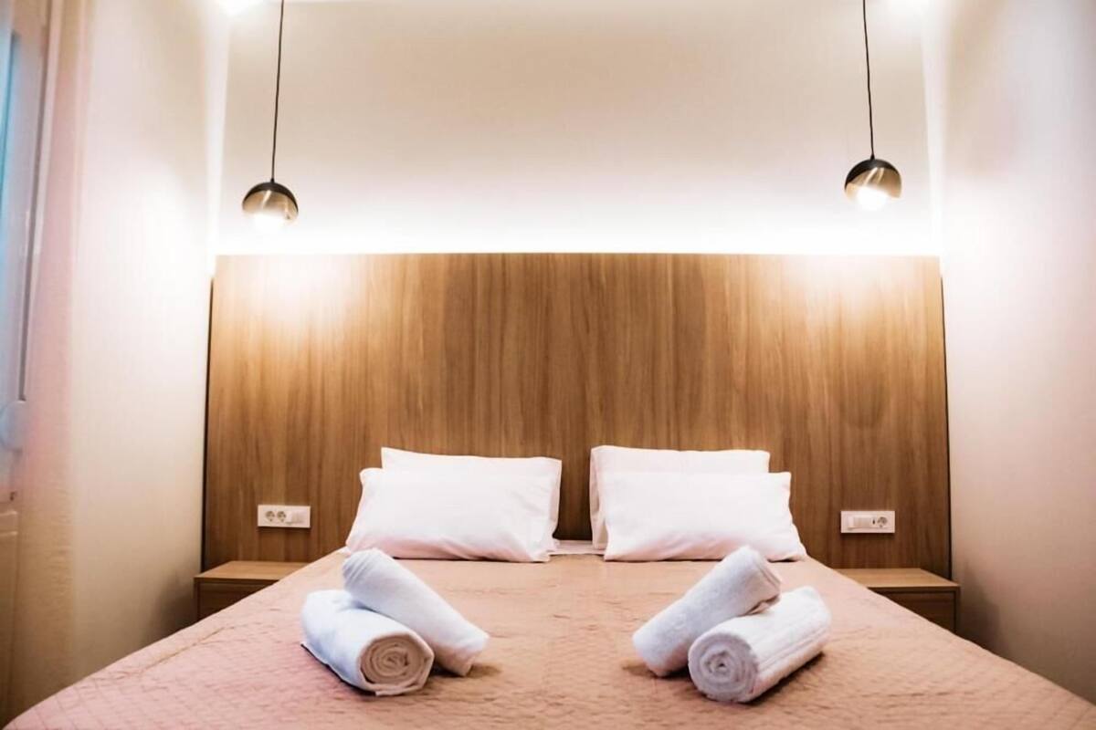 A hotel room with a wooden headboard, two white pillows, a pink bedspread, and two rolled towels on the bed.