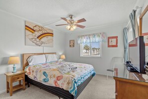 Une chambre à coucher avec un lit, un ventilateur de plafond, un bureau, une chaise et une fenêtre avec des rideaux.