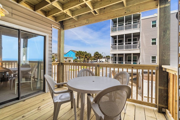 A covered outdoor area with a table and chairs, a wooden deck, and a view of a building with balconies.