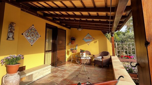 A covered patio with a wooden ceiling, yellow walls, and tiled flooring. There are wicker furniture and potted plants.