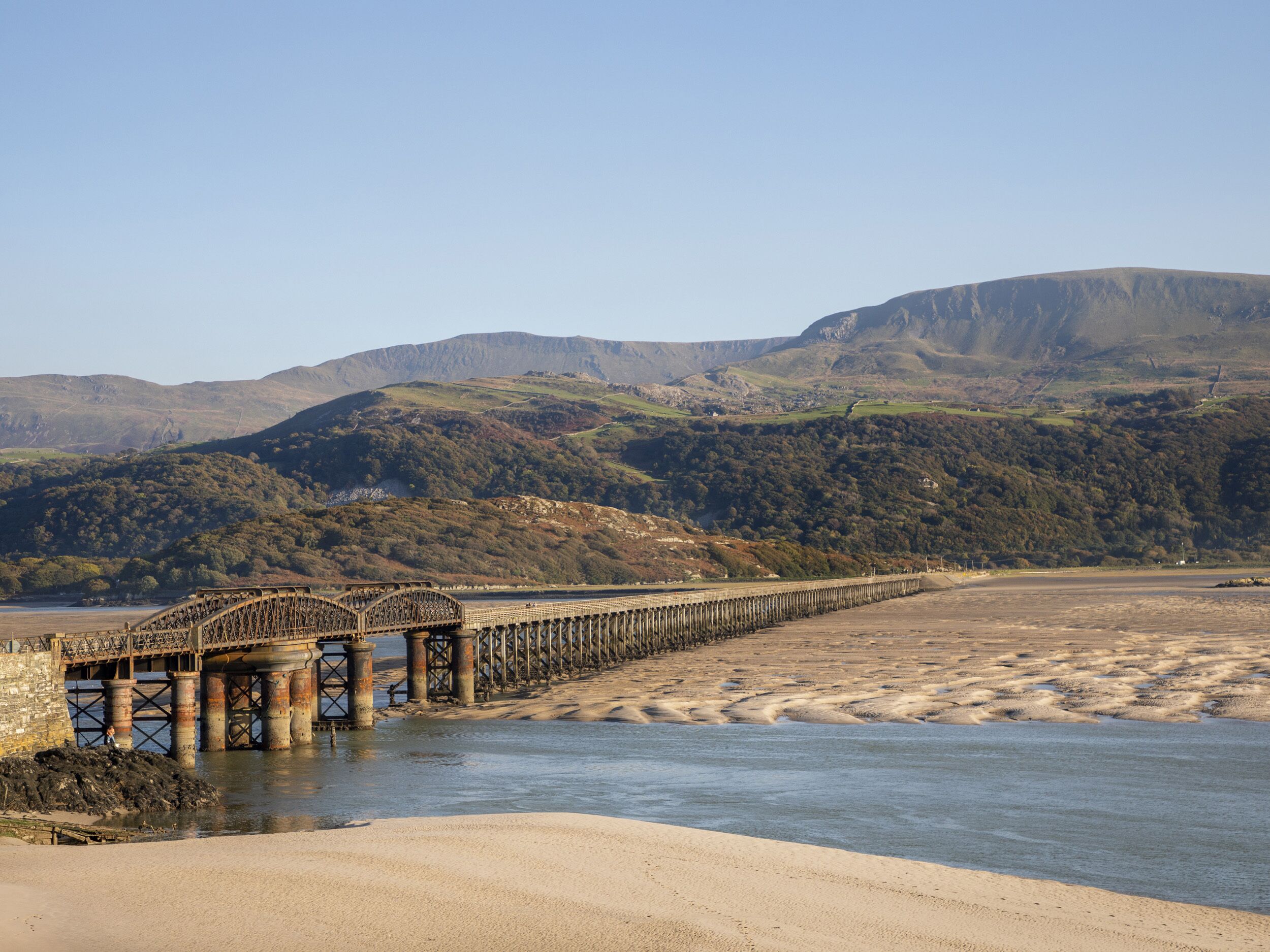 Un paisaje costero con un puente sobre el agua, una playa de arena y colinas al fondo.