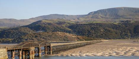 Un paisaje costero con un puente sobre el agua, una playa de arena y colinas al fondo.