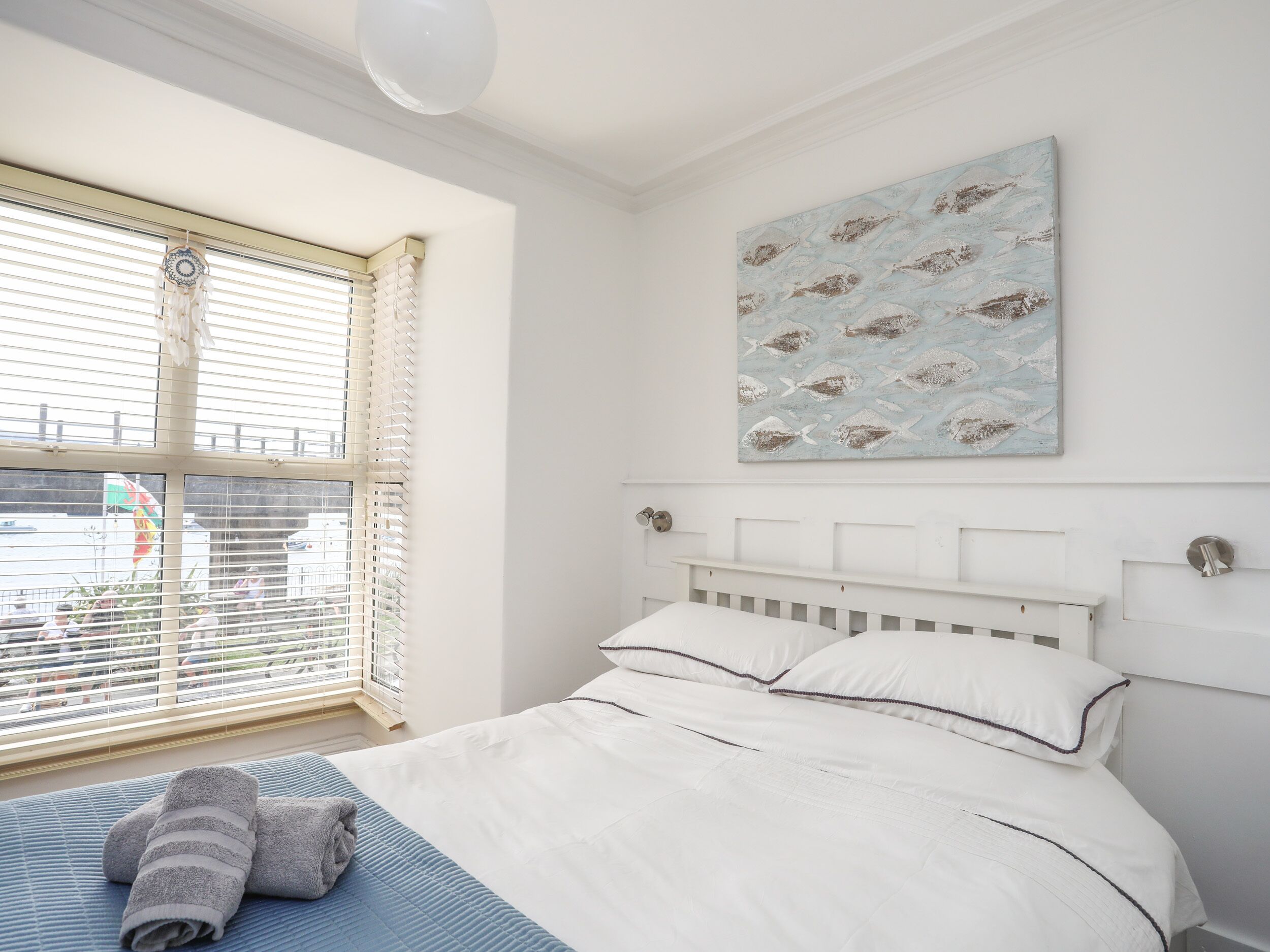 A bedroom with a bed, a window with blinds, a painting on the wall, and a view of the outside.