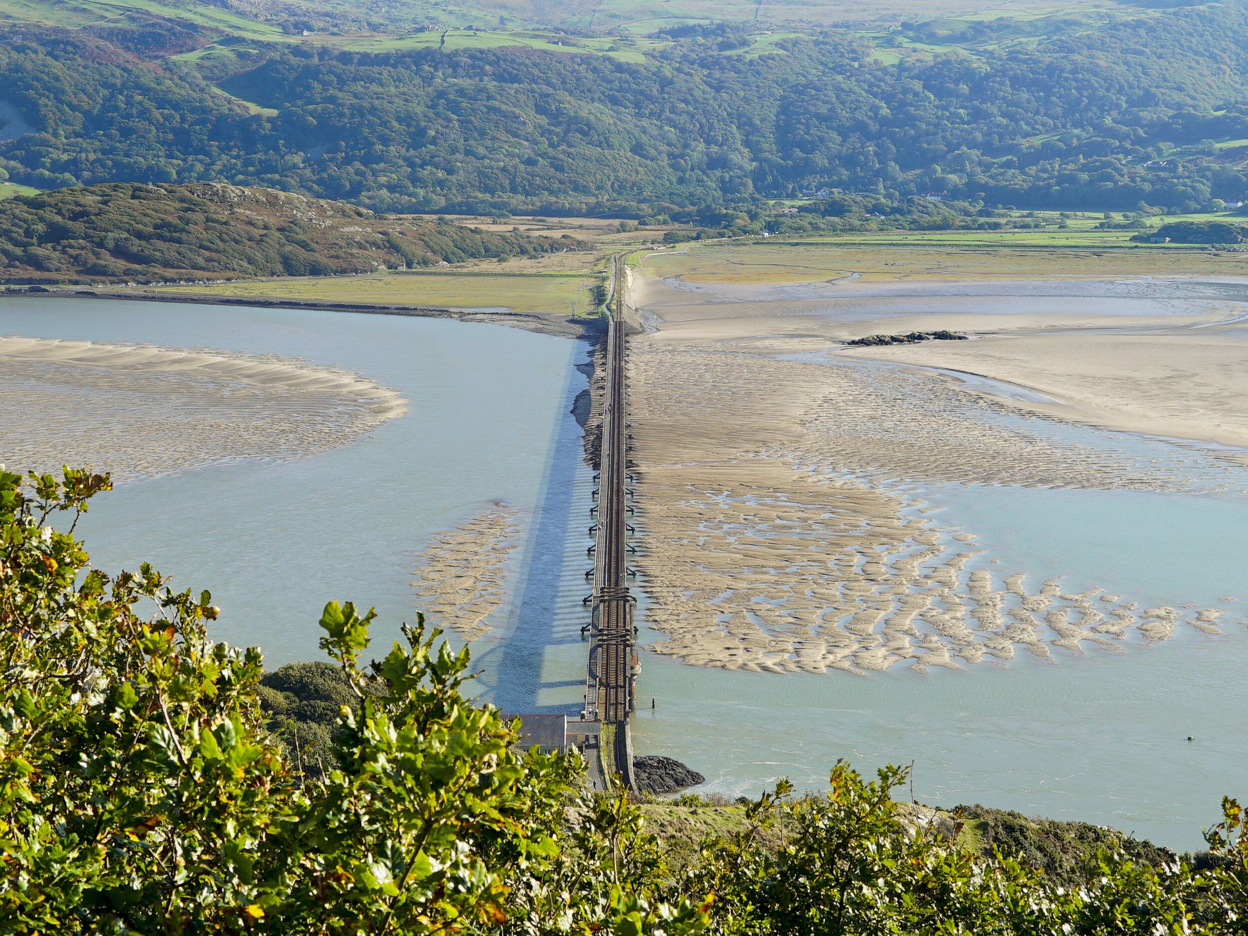 Un paysage côtier avec un pont qui s’avance dans l’eau, entouré de verdure et de collines.