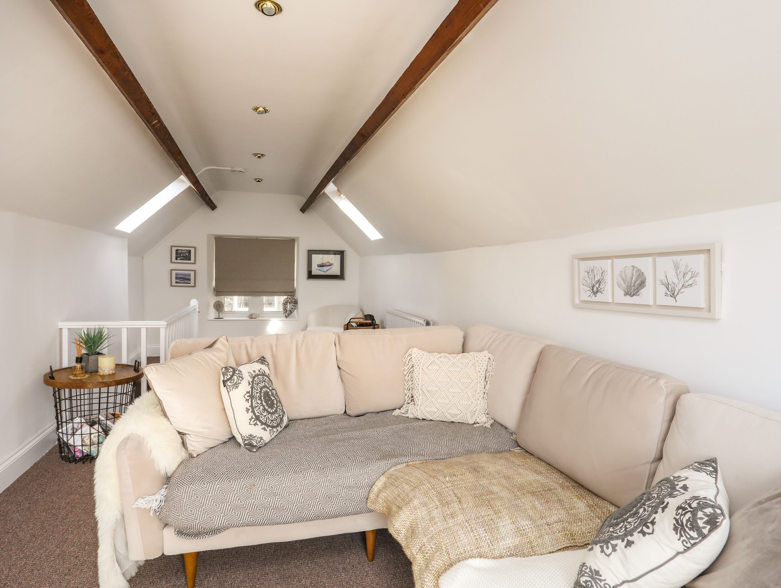 Un salon chaleureux avec un canapé d’angle beige, un plaid à motifs et un coussin blanc. On y trouve des tableaux encadrés au mur, une petite table avec une plante et un radiateur.