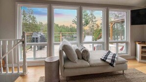 Ein Zimmer mit einer Couch, die zu großen Fenstern hin ausgerichtet ist, einer Treppe und einem Flachbildfernseher an der Wand.