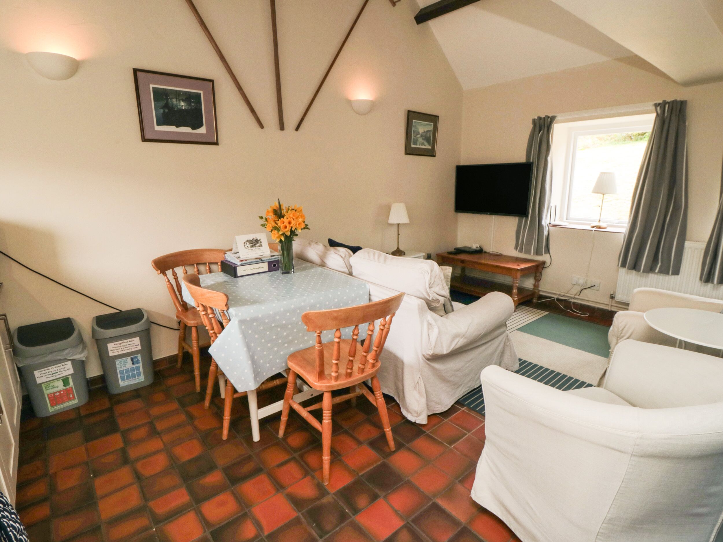 Una habitación acogedora con una mesa de comedor, sillas, un sofá y un televisor. Hay marcos de cuadros en la pared y una ventana con cortinas.