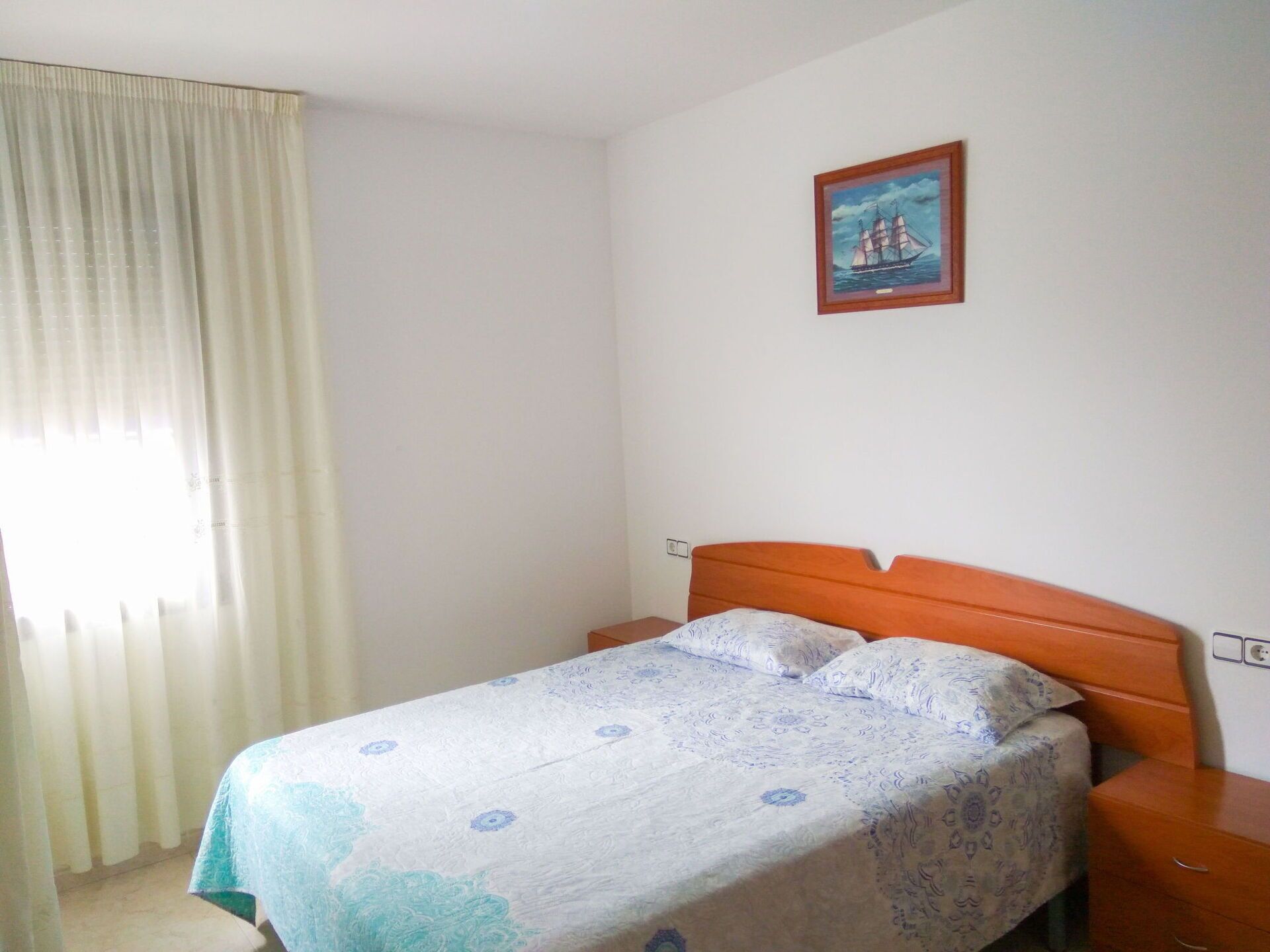 A simple bedroom with a bed, a nightstand, a wall-mounted painting, and a window with curtains.