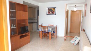 Una sala de estar con un sofá, una mesa de comedor con sillas, un televisor sobre un mueble de madera, una cocina al fondo y una puerta que da a otra habitación.