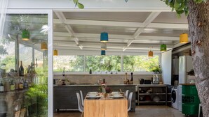 A modern kitchen with a wooden dining table, built-in appliances, and large windows offering a view of greenery.